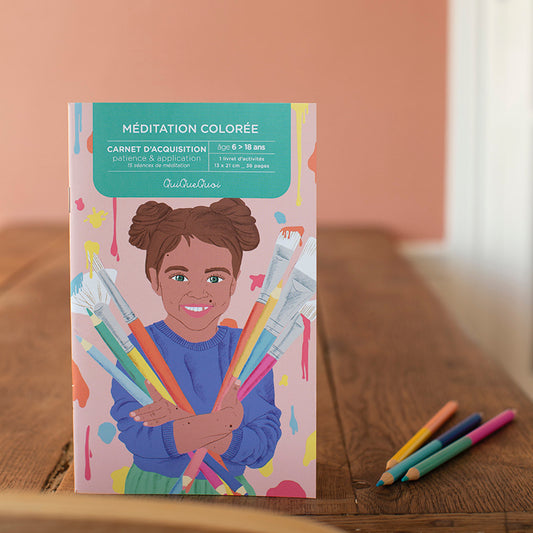 cahier de coloriages posé debout sur un bureau en bois. illustré par une petite fille joyeuse avec couettes qui porte à bras le corps des pinceaux pleins de peintures et de couleurs.