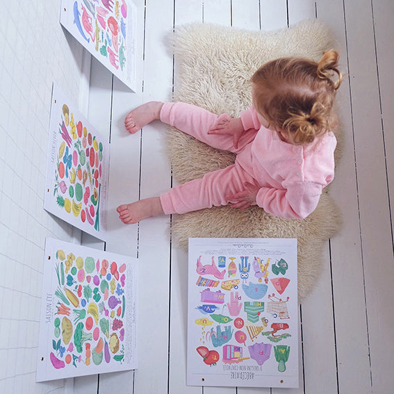 vue du dessus une petite fille en jogging rose, assise sur un tapis blanc, regarde une affiche illustrée avec des légumes et fruits de saison. Saison hiver. 