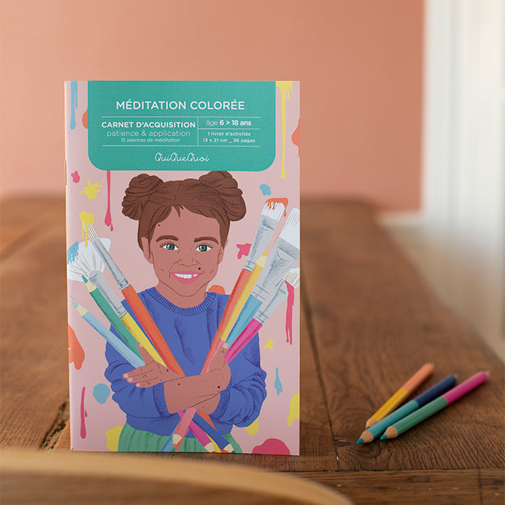 cahier de coloriages posé debout sur un bureau en bois. illustré par une petite fille joyeuse avec couettes qui porte à bras le corps des pinceaux pleins de peintures et de couleurs. 