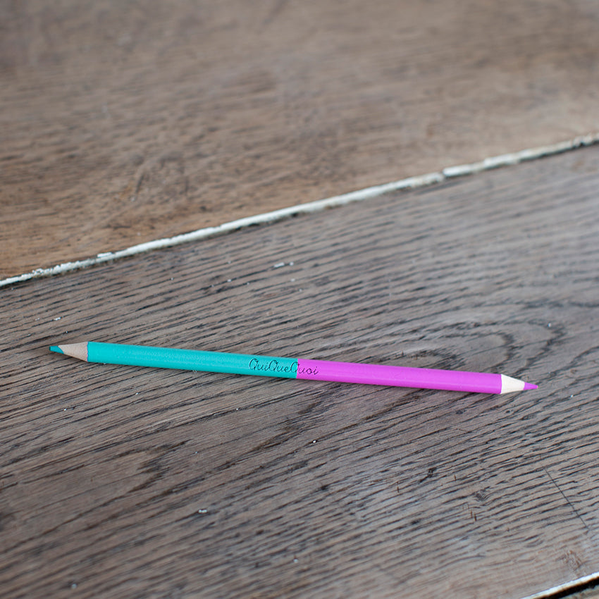 crayon bicolore avec une mine rose et une verte. Des couleurs pétillantes. Siglé en noir, au centre, QuiQueQuoi. le crayon est posé sur une table en bois