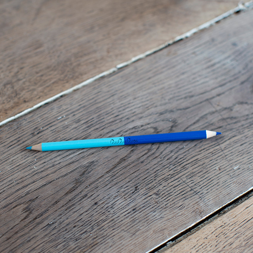 crayon bicolore avec une mine bleu clair et une bleu foncé. Des couleurs pétillantes. Siglé en noir, au centre, QuiQueQuoi. le crayon est posé sur une table en bois