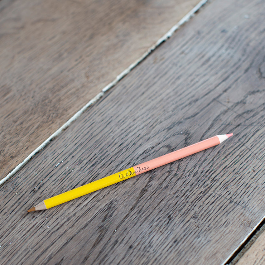 crayon bicolore avec une mine rose et une jaune. Des couleurs pétillantes. Siglé en noir, au centre, QuiQueQuoi. le crayon est posé sur une table en bois